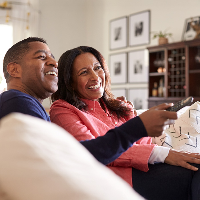 A smiling couple sits on a couch in a cozy living room. The woman, wearing a pink sweater, holds a remote control. The man, in a navy sweater, looks toward the TV, their shared laughter undeterred by his hearing loss. They appear relaxed and happy amid photos and shelves.